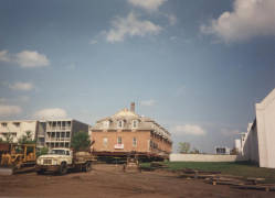 A large brick building being relocated sits on a platform above the ground. A truck and construction equipment are visible nearby. The background features clear skies and a mix of modern and older buildings.