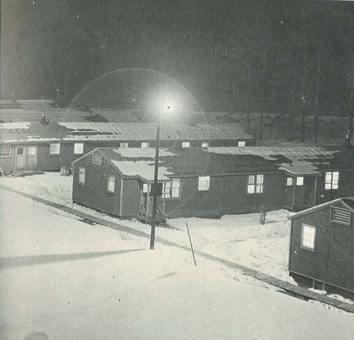 A black and white photograph shows several wooden barracks lined up in the snow. A single streetlamp illuminates the scene, casting light on the roofs and snowy ground. The setting appears quiet and remote.