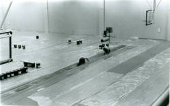 Black and white photo of two people pulling a large tarp or cover across the floor of a gymnasium. The space is mostly empty except for a few scattered items. A basketball hoop is visible on the right wall.