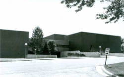 Black and white photo of a large, flat-roofed brick building with few windows. There are trees and bushes near the entrance and a street with a traffic sign in the foreground.