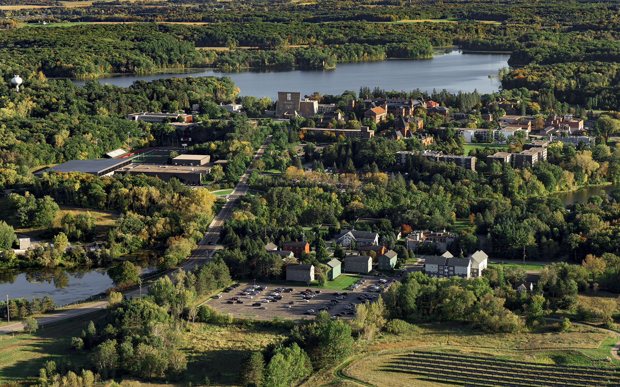 Aerial view of a campus surrounded by lush greenery and a lake. The campus features various buildings, roads, and parking lots, with expansive forests in the background.