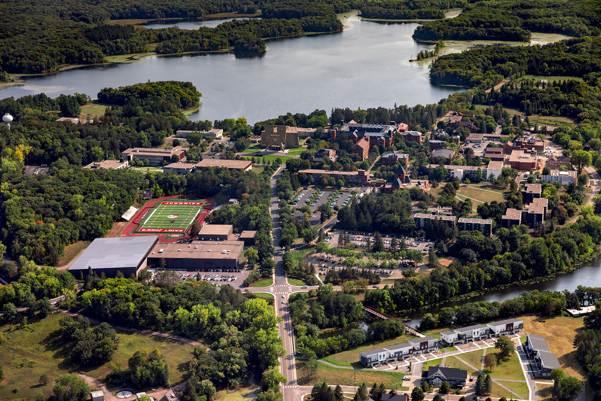 Aerial view of a lakeside campus with green sports fields, multiple buildings, and forested areas. A road bisects the scene, leading towards the water, surrounded by lush greenery and a small river.