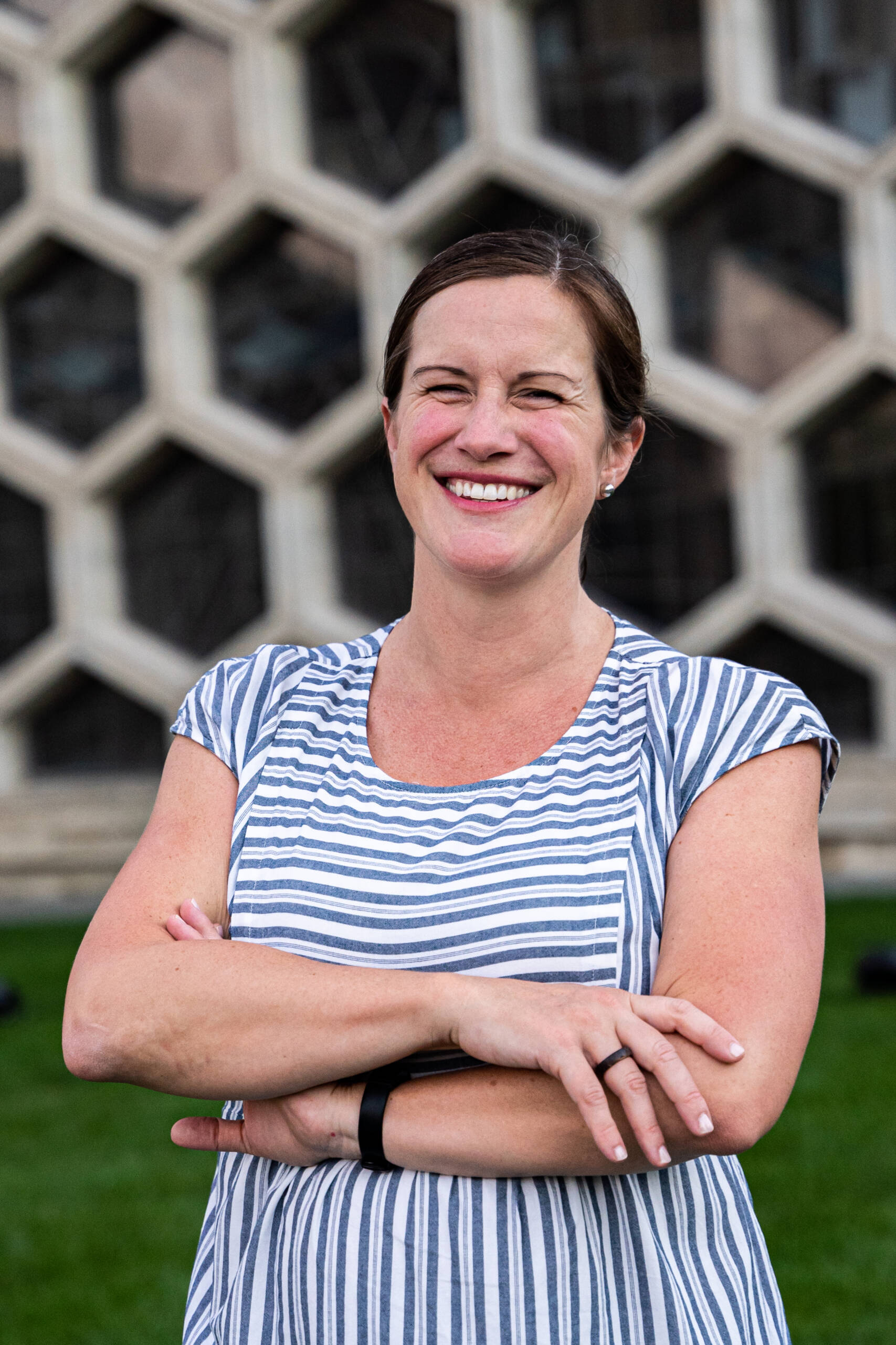 A person with a broad smile and light brown hair stands with arms crossed. They are wearing a blue and white striped shirt and an armband. The background features a building with a honeycomb-patterned facade. Green grass is visible at the bottom of the image.