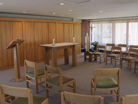 A small, simple chapel with wooden furniture. It includes an altar with two candles, a lectern with an open book, several rows of empty chairs, and a window with vertical blinds allowing natural light to enter the room.