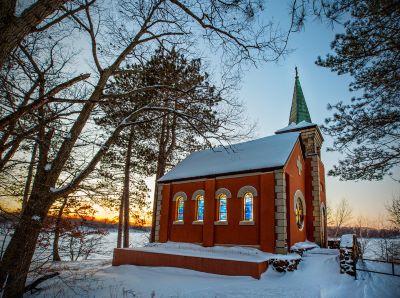 A small red-brick chapel with arched stained glass windows and a green steeple stands amidst a snowy landscape at sunset. Tall, bare trees surround the chapel, and the sky is tinged with warm hues of orange and blue.