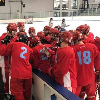 A hockey team in red jerseys with blue numbers huddles together on the ice rink bench, listening to their coach. They appear to be strategizing before a game or practice. The background shows an ice rink and a referee on the ice.
