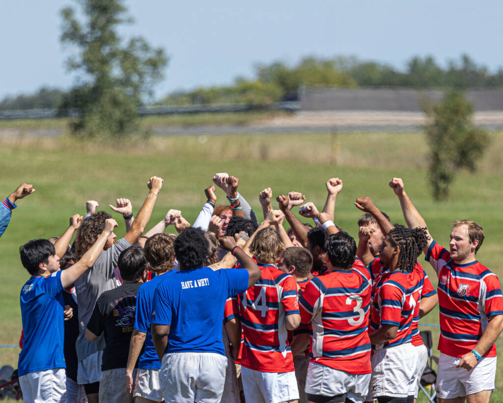 A group of rugby players, dressed in red and blue striped jerseys, huddle together with their arms raised in celebration or motivation. They are standing on a grassy field under a clear sky, with some teammates wearing blue practice shirts.
