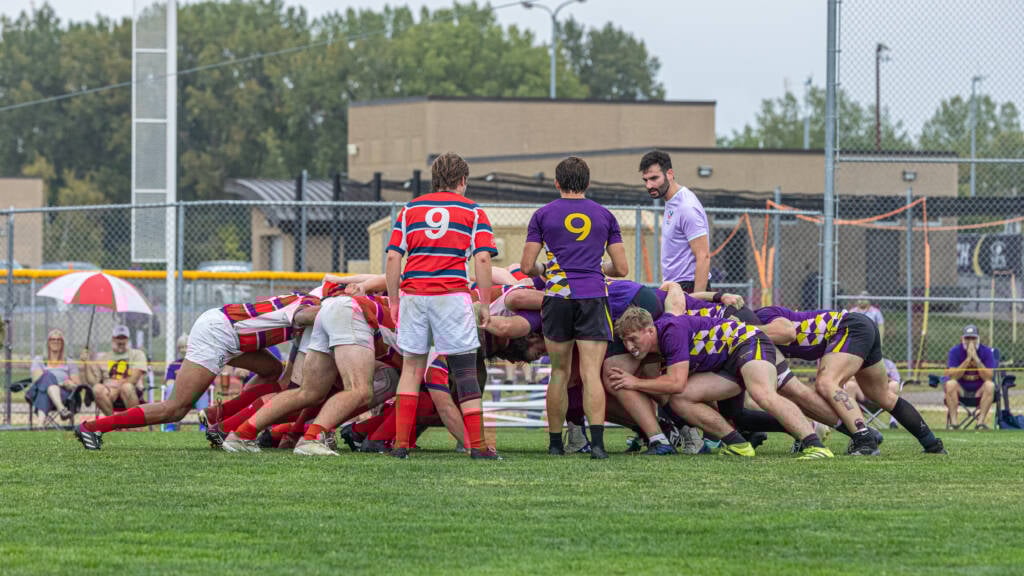 A rugby match in progress, showing two teams locked in a scrum. One team wears red and white striped jerseys, while the other wears purple jerseys with yellow checkered designs on the sides. The players are on a grassy field, and spectators are visible in the background.