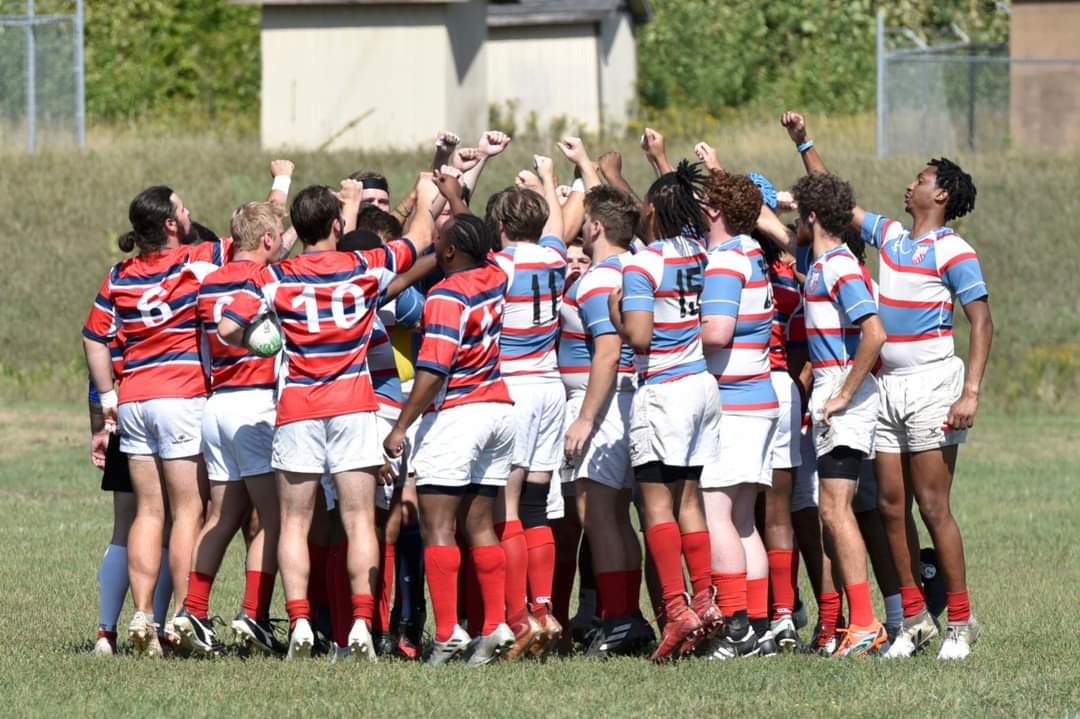 A rugby team wearing red, white, and blue uniforms, all standing in a huddle on a grassy field with their arms raised in the air. They are preparing for a match, appearing united and motivated.