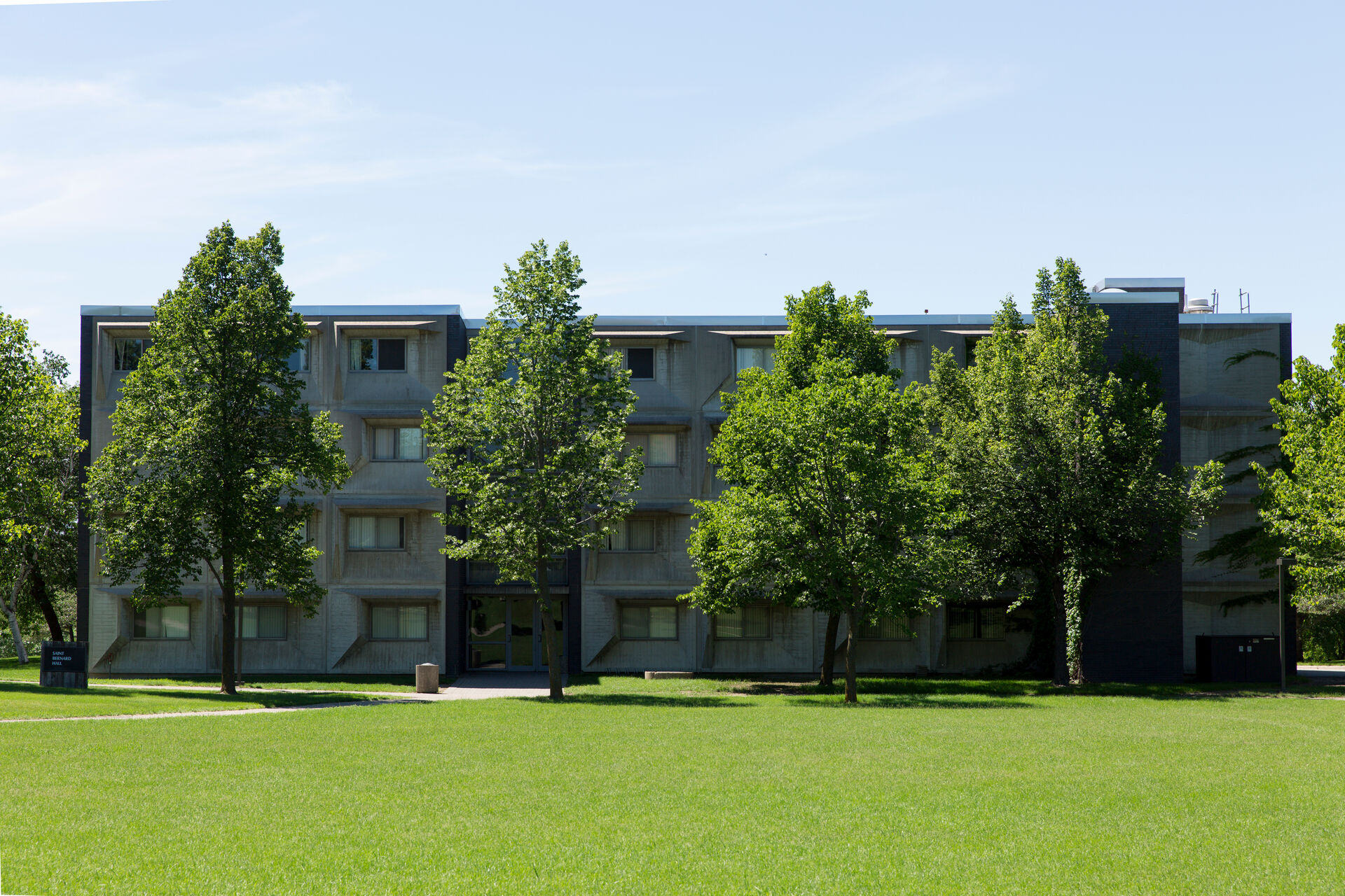 A four-story building with gray exterior and numerous windows, surrounded by lush green grass and several trees, under a clear blue sky.