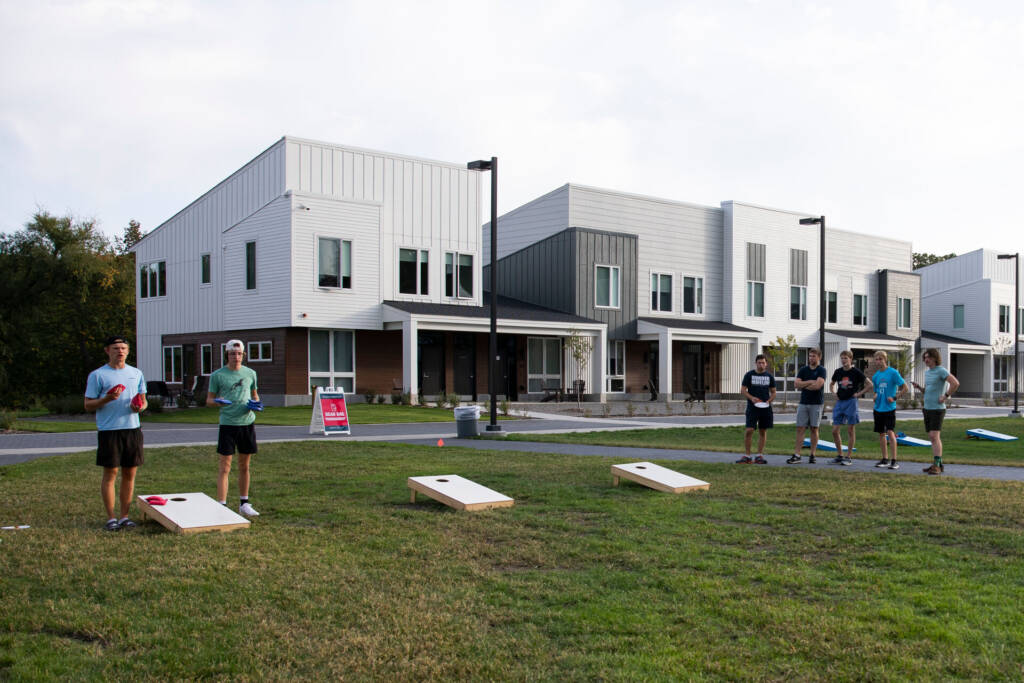 A group of people play cornhole on a grassy area in front of modern townhouses. There are six individuals, some holding bean bags, and three wooden cornhole boards arranged on the ground. The sky is slightly cloudy.