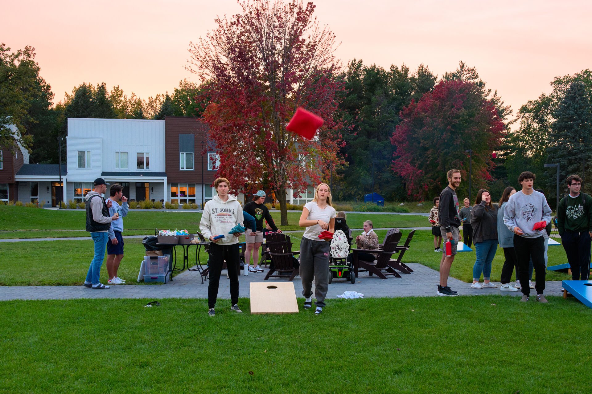 A group of people enjoying an outdoor gathering on a grassy field during sunset. Two people are playing cornhole in the foreground, while others chat and relax in the background. A mix of trees, some with autumn leaves, and modern buildings are visible.