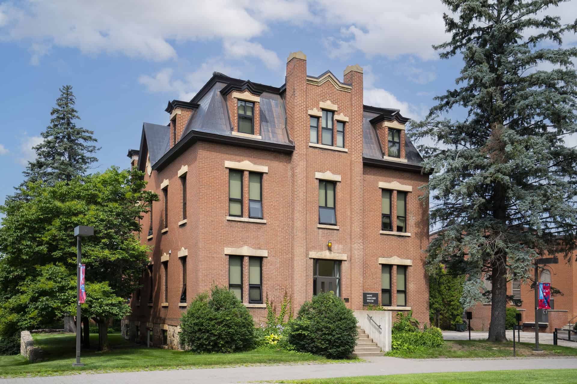 A three-story red brick building with beige trim, dormer windows, and a mansard roof, surrounded by trees and shrubs, under a blue sky with clouds.