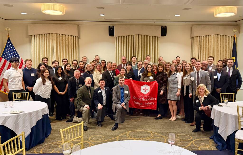 A large group of people gathered in a banquet room, posing for a group photo. Some are holding a red banner with a white emblem and text. The room features round tables with blue tablecloths and flags in the background.