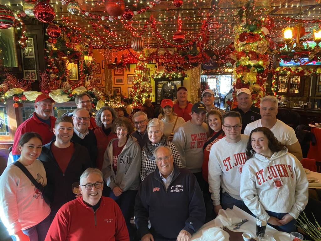 A group of people gathered at a festive, holiday-decorated venue. They are smiling and wearing casual attire, with several sporting St. John's logos on their clothing. The room is adorned with red and gold ornaments and twinkling lights.