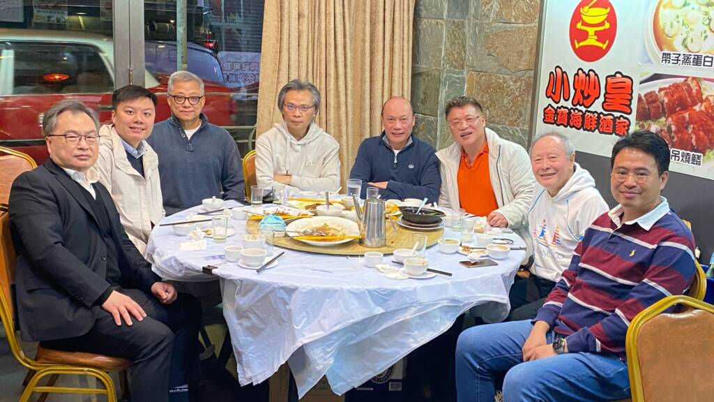 Eight men sit around a round table in a restaurant, partially covered with a white cloth. They are smiling and have finished a meal together. The restaurant wall features a red and yellow logo with Chinese text.