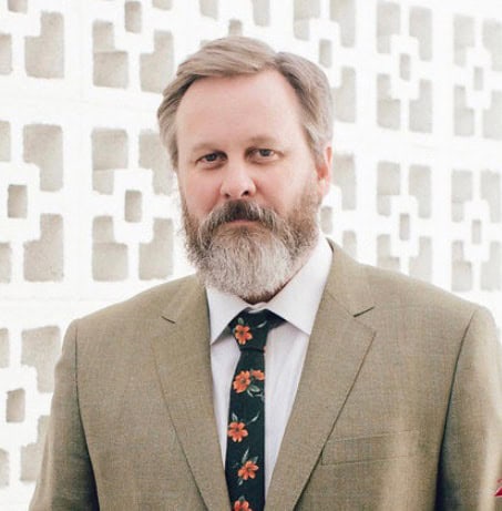 A man with a gray beard and mustache wearing a light brown suit and a floral tie stands in front of a white patterned wall.