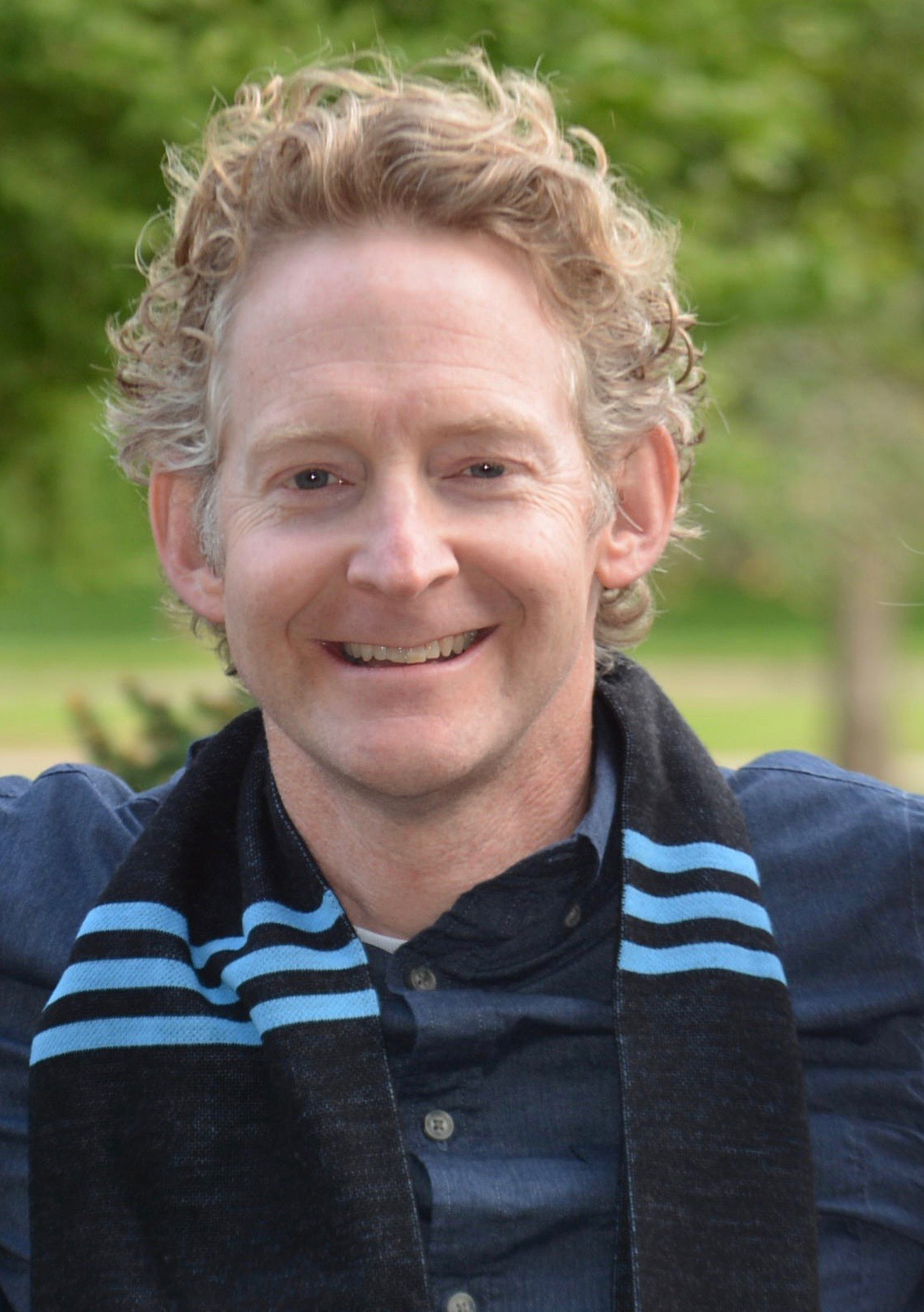 A smiling man with curly light hair wearing a navy shirt and a dark scarf with light blue stripes stands outdoors with greenery in the background.
