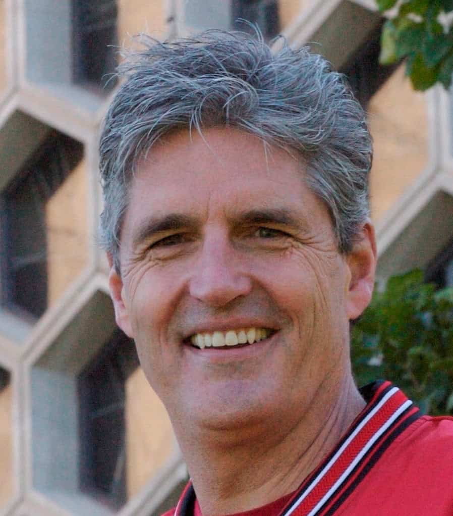 A middle-aged man with gray hair smiles outdoors. He is wearing a red shirt and a jacket, with a geometric patterned building and green leaves visible in the background.