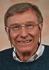 A smiling older man with short light brown hair, glasses, and a blue sweater over a collared shirt, posing in front of a neutral background.