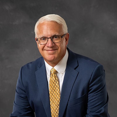 A middle-aged man with short white hair and glasses, wearing a navy blue suit, white shirt, and yellow patterned tie, poses in front of a dark gray background, smiling slightly.
