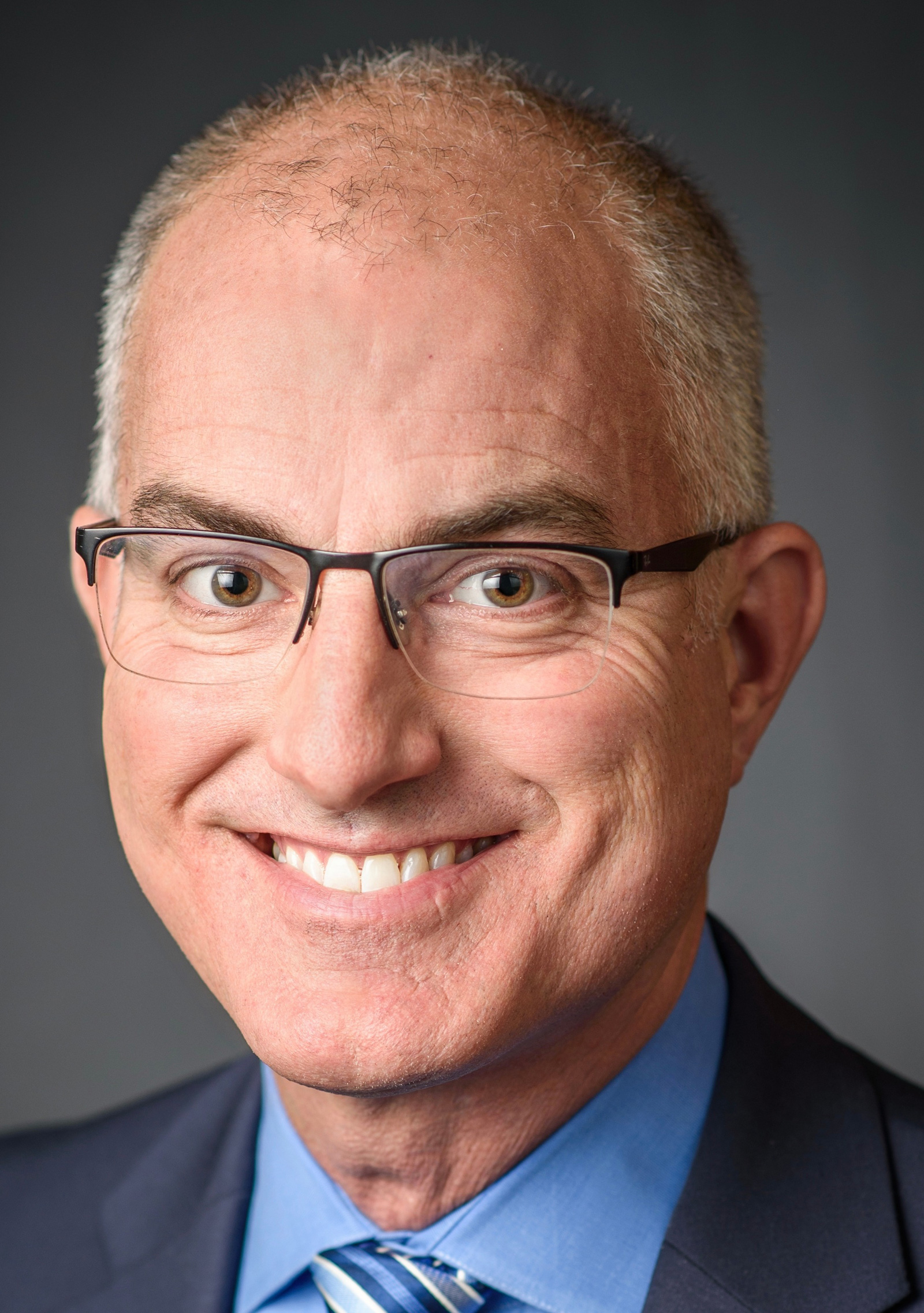 A middle-aged man with short gray hair and glasses, wearing a blue dress shirt, striped tie, and dark suit jacket, smiles at the camera against a dark background.