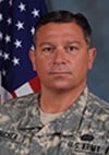 A man in a U.S. Army uniform poses for an official portrait in front of an American flag and a gray background.