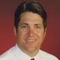 A man with short dark hair is smiling, wearing a white shirt and a red tie, pictured against a solid dark red background.