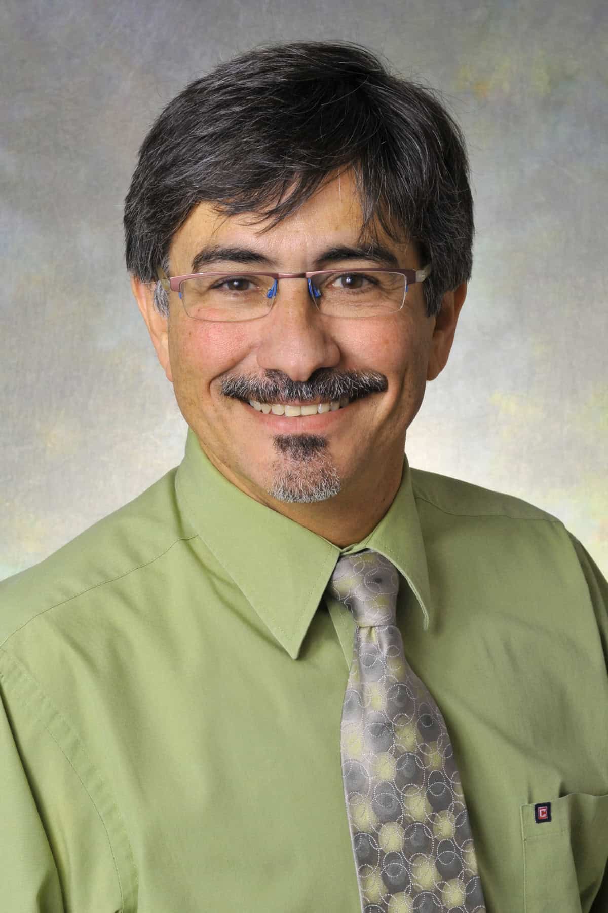 A middle-aged man with short dark hair, glasses, and a mustache smiles at the camera. He is wearing a light green dress shirt and a patterned gray tie, posed in front of a neutral, softly colored background.