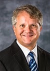 A man with short, wavy gray hair, wearing a dark pinstripe suit, white shirt, and blue striped tie, smiling in front of a blurred gray and blue background.