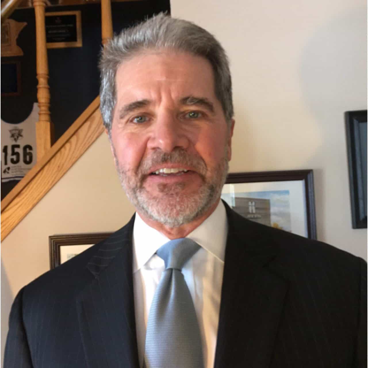 A middle-aged man with gray hair and a beard, wearing a dark pinstripe suit, white shirt, and light blue tie, stands indoors in front of framed pictures and a staircase.