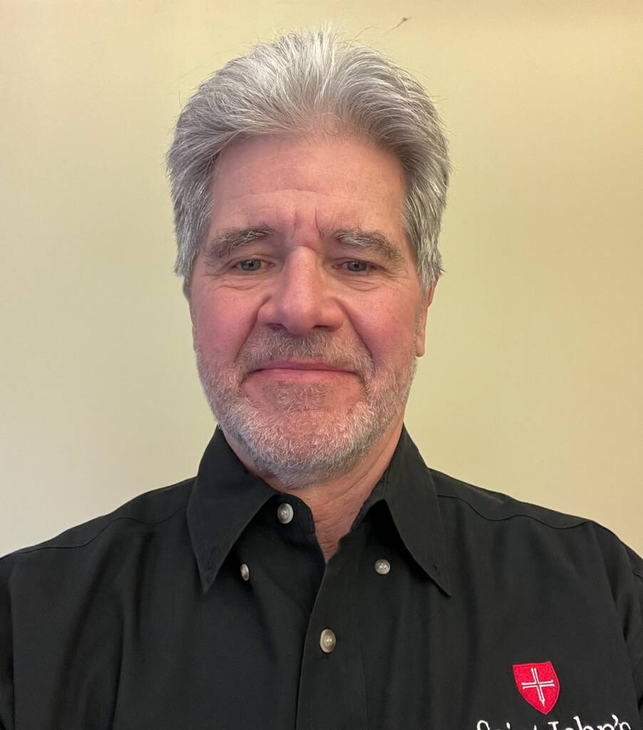 A middle-aged man with gray hair and a beard smiles at the camera. He is wearing a black collared shirt with a red and white crest logo and the words "Saint John's" visible on the shirt. The background is plain beige.