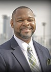 A man in a dark suit, white shirt, and plaid tie smiles at the camera. He has a short beard and closely cropped hair. The background shows a blurred cityscape with tall buildings.