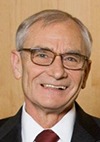 An older man with gray hair and glasses, wearing a dark suit, white shirt, and red tie, smiles in front of a light wood background.