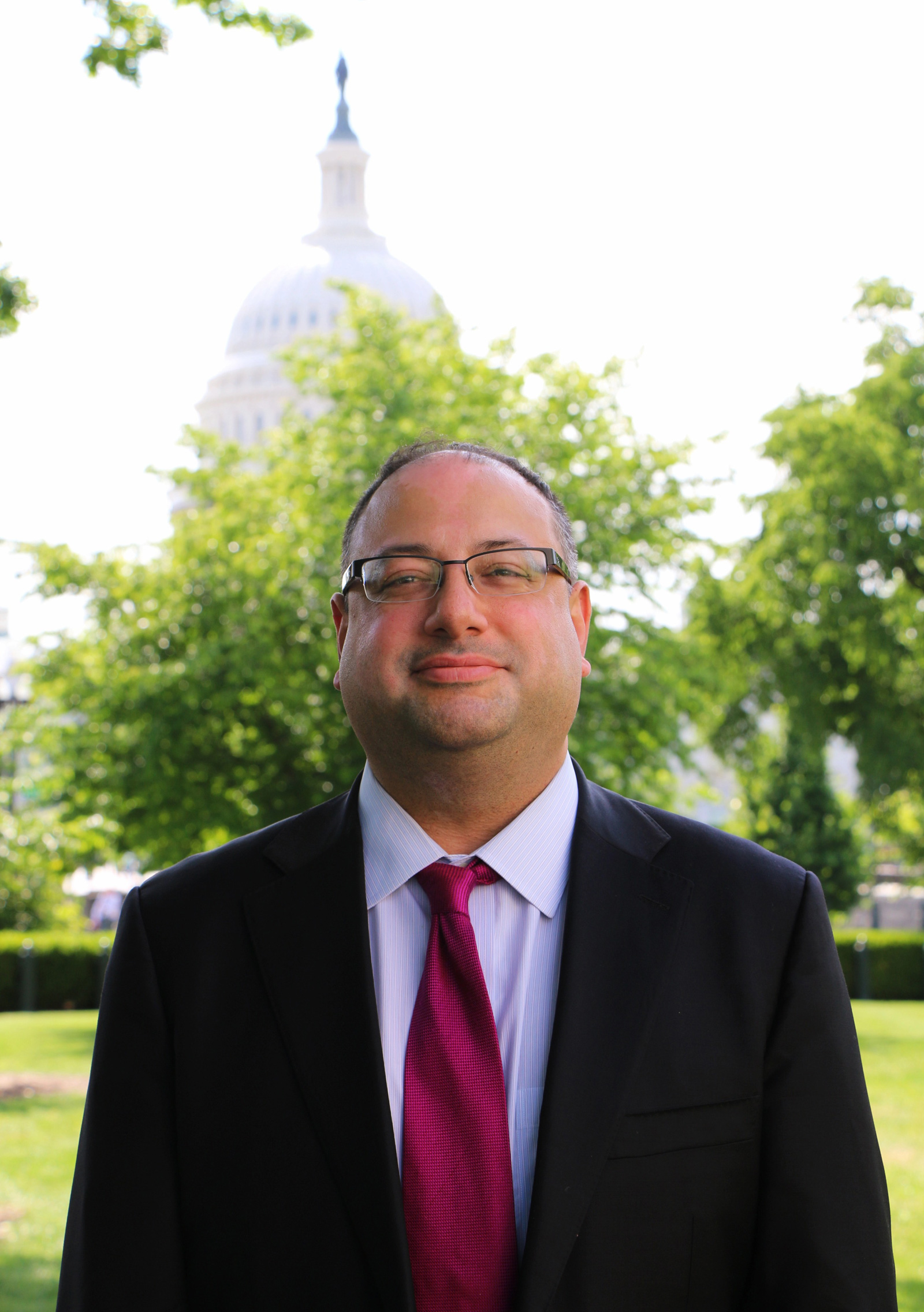 A man in a suit and red tie stands outside, smiling, with trees and the dome of the U.S. Capitol building visible in the background.