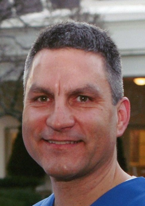 A middle-aged man with short dark hair and wearing a blue shirt smiles at the camera outdoors, with blurred buildings and trees in the background.