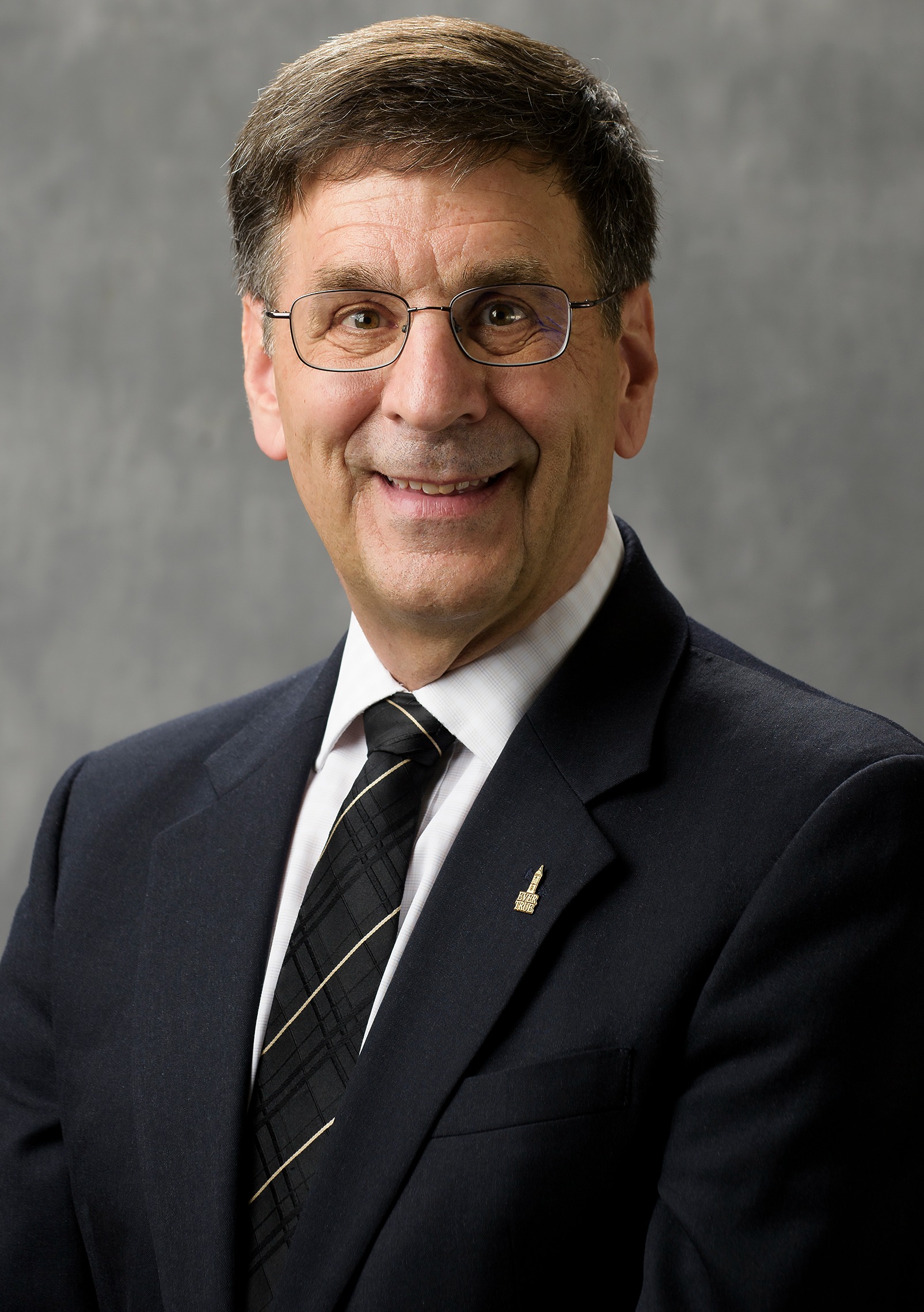 A middle-aged man with short dark hair and glasses, wearing a dark suit, white shirt, and striped tie, smiles at the camera against a plain gray background.