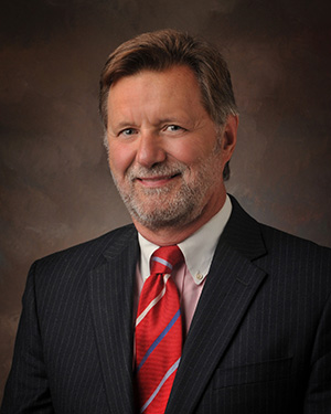 A middle-aged man with light brown hair and a beard, wearing a dark pinstripe suit, white shirt, and red striped tie, poses smiling against a dark, textured studio background.