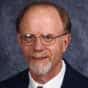 A middle-aged man with glasses, a short beard, and balding hair, wearing a suit and tie, poses in front of a blue-gray mottled background.