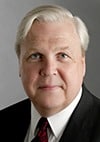 A middle-aged man with light skin, short white hair, and blue eyes is wearing a dark suit, white shirt, and patterned tie. He is looking directly at the camera against a plain gray background.