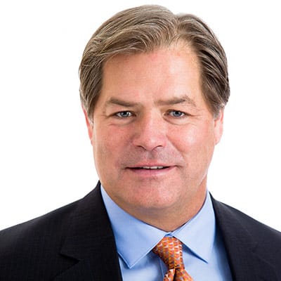 A middle-aged man with short, light brown hair wearing a dark suit, light blue shirt, and patterned orange tie, smiling slightly in front of a plain white background.