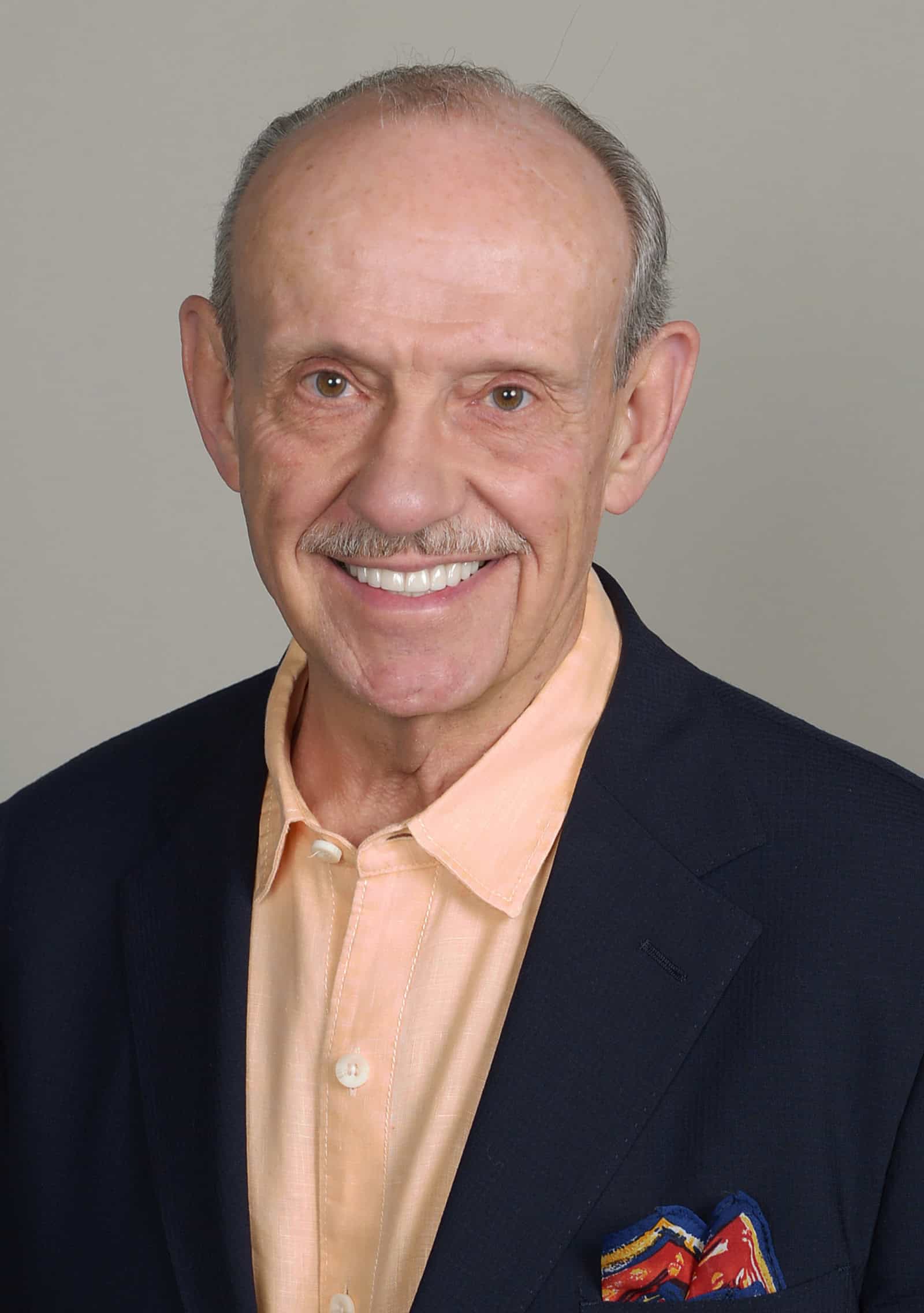 An older man with a receding hairline, wearing a navy blazer over a peach button-up shirt, smiles at the camera against a plain, light gray background.