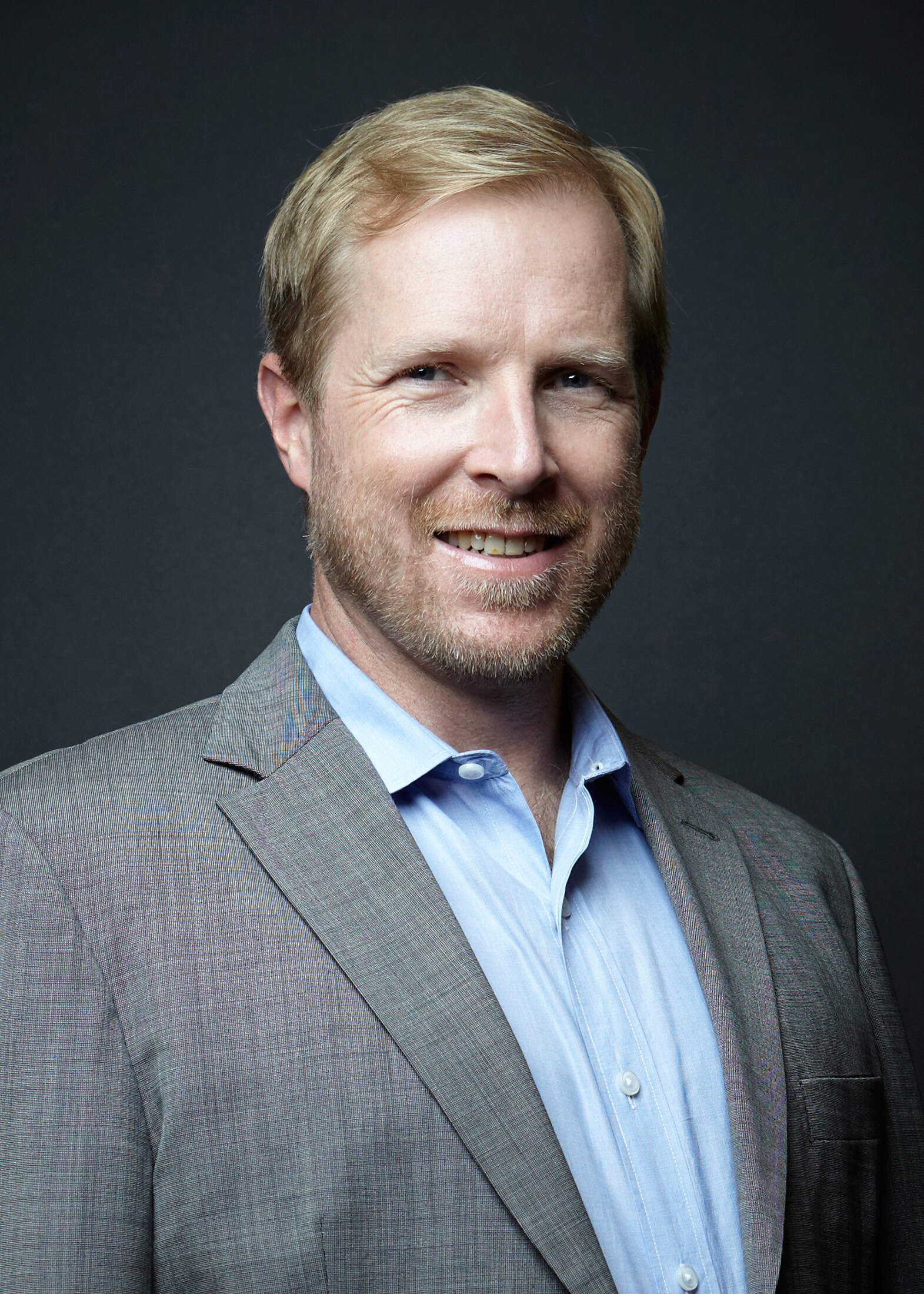 A man with short blond hair and a trimmed beard, wearing a light blue dress shirt and a gray suit jacket, smiles at the camera against a dark background.