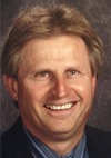 A man with light brown hair and blue eyes smiles at the camera. He is wearing a dark blazer over a blue collared shirt and tie, with a neutral, softly lit background behind him.