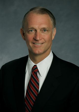 A middle-aged man wearing a dark suit, white shirt, and red striped tie, smiling at the camera against a plain grey background.