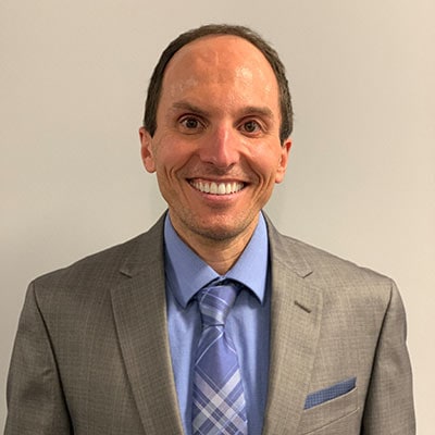 A man wearing a light gray suit, blue dress shirt, and plaid tie smiles at the camera while standing against a plain light-colored background.