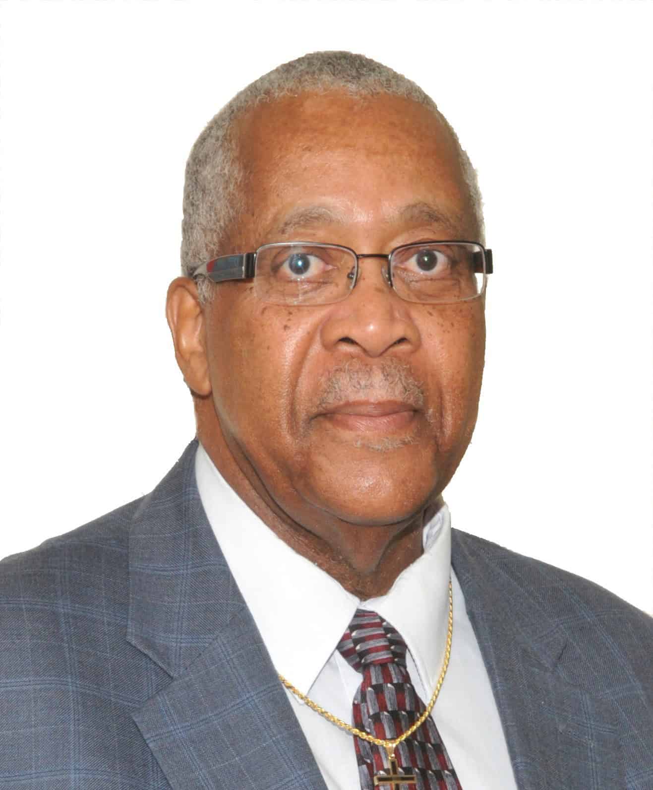 An older man with short gray hair and glasses wearing a blue plaid suit, white shirt, patterned tie, and a gold necklace, posing against a plain white background.
