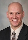 A man with a bald head, wearing a dark suit, light shirt, and patterned tie, smiling at the camera against a neutral background.