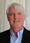 A man with short white hair is smiling, wearing a light blue collared shirt and a dark blazer, standing in front of white curtains.