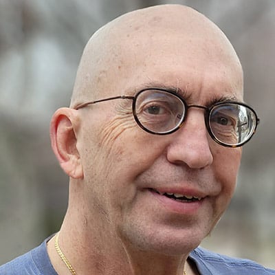 A bald man with round glasses and a gold chain smiles slightly at the camera. He is wearing a blue shirt, and the background is blurred, showing an outdoor setting.